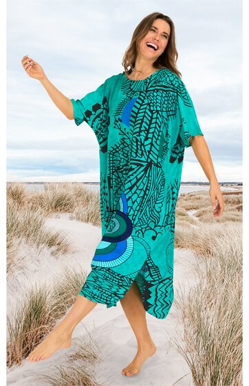 Kleid in türkis mit abstraktem dunkelblauem Muster, Rundhalsausschnitt, halblange lockere Ärmel, knielang, Figur umspielend, getragen von lachender Frau barfuß, stehend auf Sand mit Strandgras
