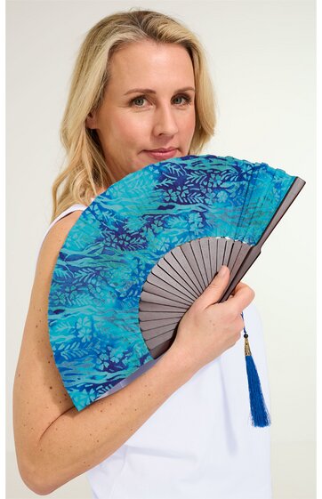 Handfächer in Türkis mit dunkelblauem floralem Muster, grauer Fächergriff mit blauer Quaste, gehalten von stehender Frau in weißem ärmellosem Oberteil