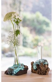 Zwei Vasen aus transparentem Glas mit unregelmäßigen Rändern, auf braunem Treibholz befestigt, eine Vase mit hoher schmaler Form und Pflanze mit weißen Blüten, andere kurze Vase ohne Inhalt, auf hellbraunem Holztisch vor unscharfem Hintergrund