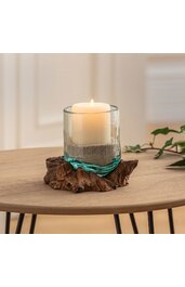Kerze in glaszylindrischer Form, durchscheinendes türkisfarbenes Glas mit Sandfüllung, auf unregelmäßig geformtem braunem Holzsockel, brennende weiße Stumpenkerze, dekoratives Wohnaccessoire, auf rundem Holztisch