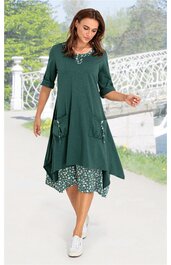 Grünes Zweiteiler-Kleid mit asymmetrischem Schnitt, Oberteil ohne Muster, halbärmlig, knielang, mit Taschen und Knopfdetails, darunter floral gemustertes Kleid in Grün- und Weißtönen, getragen von stehender Frau in weißer Freizeitschuhe