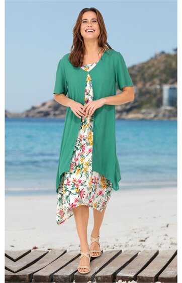 Zweiteiliges Sommerkleid mit hellgrünem, locker fallendem Überwurf, bunt gemustertes Blumenkleid darunter, knielang, rundhals, kurze Ärmel, getragen von lachender Frau stehend am Strand mit Meer und bewaldetem Hang im Hintergrund