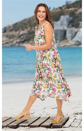 Weißes Midikleid mit buntem Blumenmuster, ärmellos, V-Ausschnitt, locker sitzend, getragen von stehender Frau am Strand, ausgestellter Rock, seitliche Taschen