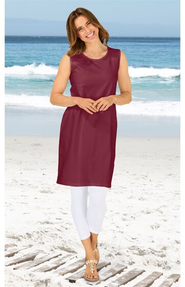Ärmelloses, knielanges Kleid in dunkelrot, ohne Muster, rundhalsausschnitt, gerade geschnitten, getragen von lächelnder stehender Frau am Strand, kombiniert mit weißen Leggings und Sandalen