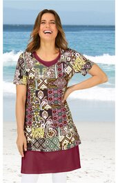 Kurzärmeliges Kleid in Burgunderrot und gemischtem Ethno-Muster mit Quadraten und Kreisen in Beige, Grün, Braun und Gelb, Rundhalsausschnitt, locker sitzend, getragen von stehender lachender Frau am Strand
