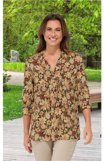 Locker geschnittene Bluse in Braun mit floralen Mustern in Gelb und Rot, V-Ausschnitt, lange Ärmel mit Bündchen, getragen von stehender Frau vor grünem Hintergrund