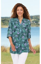 Damenbluse, grün mit abstraktem blauen und lila Muster, V-Ausschnitt mit Stehkragen, lange Ärmel, locker sitzend, getragen von stehender Frau vor Wasserkulisse