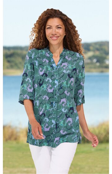Damenbluse, grün mit abstraktem blauen und lila Muster, V-Ausschnitt mit Stehkragen, lange Ärmel, locker sitzend, getragen von stehender Frau vor Wasserkulisse