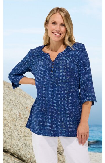 Damenbluse in Dunkelblau mit kleinen weißen Punkten, V-Ausschnitt mit Knopfleiste, 3/4-Ärmel, locker sitzend, getragen von lächelnder Frau stehend vor Felsen und Meer