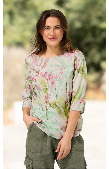 Bluse in hellem Pastellgrün mit floralem Muster in Rosatönen und zarten Grüntönen, rundhalsausschnitt, dreiviertellange Ärmel, lockere Passform, getragen von stehender Frau mit Händen in Taschen