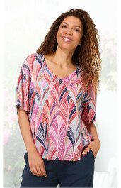 Kurzärmelige Bluse in Weiß mit buntem Blättermuster in Pink, Rot, Blau und Lila, V-Ausschnitt, lockere Passform, getragen von stehender Frau mit Händen in den Hosentaschen