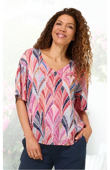 Kurzärmelige Bluse in Weiß mit buntem Blättermuster in Pink, Rot, Blau und Lila, V-Ausschnitt, lockere Passform, getragen von stehender Frau mit Händen in den Hosentaschen