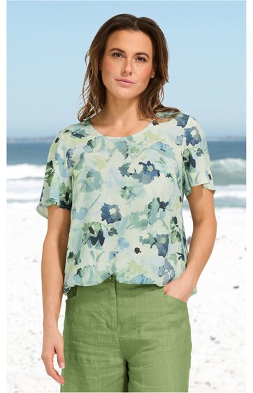 Bluse in hellem Mintgrün mit blauem Blumenmuster, Rundhalsausschnitt, kurze Ärmel, lockere Passform, getragen von woman vor Strandozean im Hintergrund