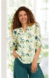 Bluse in Cremeweiß mit grünem und gelbem Blumenmuster, V-Ausschnitt mit Schlitz und Knopf, 3/4-Arm, locker sitzend, getragen von stehender Frau mit lockigem braunem Haar, grüne Hose