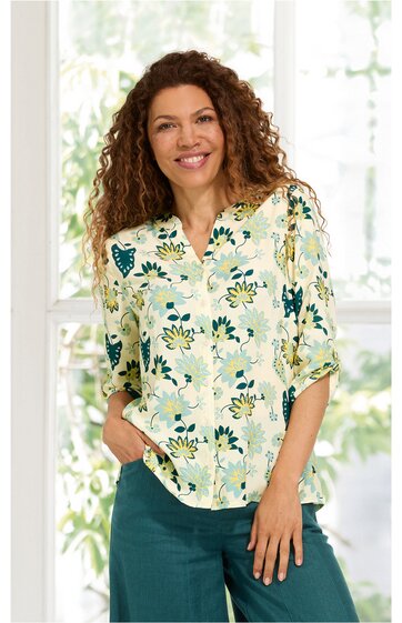 Bluse in Cremeweiß mit grünem und gelbem Blumenmuster, V-Ausschnitt mit Schlitz und Knopf, 3/4-Arm, locker sitzend, getragen von stehender Frau mit lockigem braunem Haar, grüne Hose