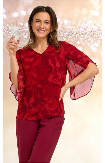 Rotes Damenoberteil mit abstraktem Muster, V-Ausschnitt, weite kurze Ärmel mit transparenten Einsätzen, tailliert, getragen von lächelnder Frau mit hochgezogenem Arm und Glas in der Hand