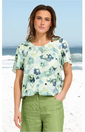 Bluse in hellem Mintgrün mit blauem Blumenmuster, Rundhalsausschnitt, kurze Ärmel, lockere Passform, getragen von woman vor Strandozean im Hintergrund