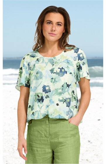 Bluse in hellem Mintgrün mit blauem Blumenmuster, Rundhalsausschnitt, kurze Ärmel, lockere Passform, getragen von woman vor Strandozean im Hintergrund