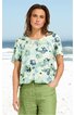Bluse in hellem Mintgrün mit blauem Blumenmuster, Rundhalsausschnitt, kurze Ärmel, lockere Passform, getragen von woman vor Strandozean im Hintergrund