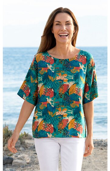 Blusenshirt türkis mit großem floralen Blatt- und Blumenmuster in Orange, Rot und Hellgrün, kurze, weite Ärmel, runder Ausschnitt, gerade geschnitten, getragen von stehender Frau vor Meereskulisse