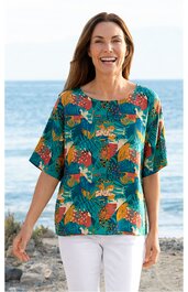 Blusenshirt türkis mit großem floralen Blatt- und Blumenmuster in Orange, Rot und Hellgrün, kurze, weite Ärmel, runder Ausschnitt, gerade geschnitten, getragen von stehender Frau vor Meereskulisse