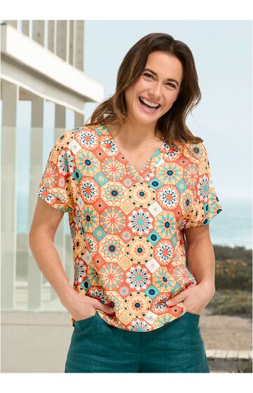 Bunte Damenbluse in Orange mit Blumen- und Kreismuster, V-Ausschnitt, kurze Ärmel, locker sitzend, getragen von lächelnder stehender Frau mit Händen in den Hosentaschen