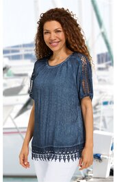 Marineblaue Bluse mit halblangen Ärmeln, Häkelspitzen-Details an Ärmeln und Saum, rundem Halsausschnitt, locker sitzend, getragen von lächelnder Frau mit lockigen dunklen Haaren, stehend vor maritimem Hintergrund