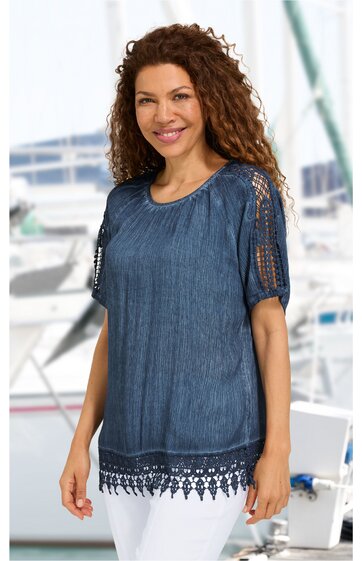 Marineblaue Bluse mit halblangen Ärmeln, Häkelspitzen-Details an Ärmeln und Saum, rundem Halsausschnitt, locker sitzend, getragen von lächelnder Frau mit lockigen dunklen Haaren, stehend vor maritimem Hintergrund