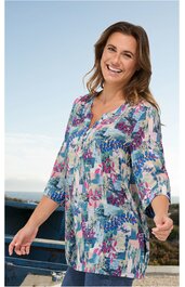 Tunika für Damen, mehrfarbig mit floralem Muster in Blau-, Rosa- und Grüntönen, langer Schnitt, Knopfleiste am Ausschnitt, 3/4-Ärmel, getragen von stehender lächelnder Frau