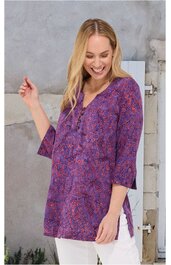 Lila Tunika mit floralem Muster in Pink und Blau, dreiviertelärmel, V-Ausschnitt mit Knopfleiste, seitliche Schlitze, locker sitzend, getragen von lächelnder Frau mit blondem, schulterlangem Haar, stehende Pose