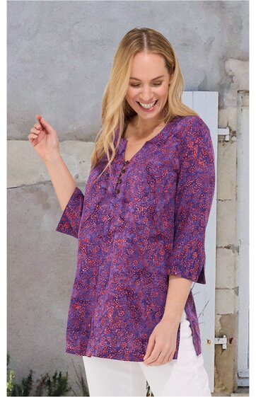 Lila Tunika mit floralem Muster in Pink und Blau, dreiviertelärmel, V-Ausschnitt mit Knopfleiste, seitliche Schlitze, locker sitzend, getragen von lächelnder Frau mit blondem, schulterlangem Haar, stehende Pose