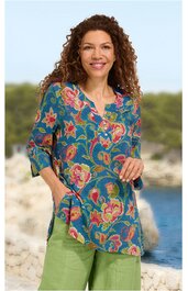 Tunikenbluse in blau mit großem floralen Muster in Rot und Grün, V-Ausschnitt ohne Kragen, 3/4-Ärmel, seitliche Taschen, locker sitzend, getragen von stehender Frau vor Küstenlandschaft