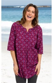 Damenbluse in purpur mit blau-weißem Blumenmuster, V-förmiger Ausschnitt ohne Kragen, dreiviertellange Ärmel, locker sitzend, getragen von stehender Frau am Strand