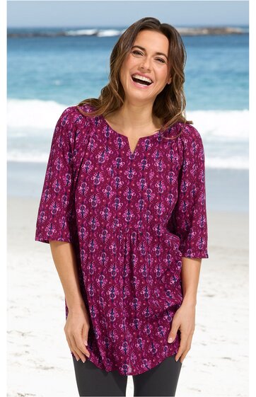 Damenbluse in purpur mit blau-weißem Blumenmuster, V-förmiger Ausschnitt ohne Kragen, dreiviertellange Ärmel, locker sitzend, getragen von stehender Frau am Strand