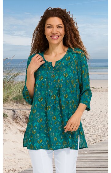 Tunika in türkisgrün mit gelb-blauem Blattmuster, V-Ausschnitt mit Knopfleiste, 3/4-ärmel, seitliche Schlitze, lockere Passform, getragen von lächelnder Frau mit lockigem braunem Haar, Strand im Hintergrund