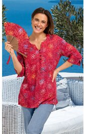 Rotes Blusenshirt mit runden weißen und orangefarbenen Mustern, V-Ausschnitt mit Knopfleiste, dreiviertellange Ärmel, locker geschnitten, getragen von lächelnder stehender Frau mit Fächer in der rechten Hand vor Außenbereich mit Korbmöbeln und Pflanz