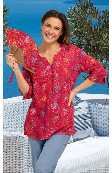 Rotes Blusenshirt mit runden weißen und orangefarbenen Mustern, V-Ausschnitt mit Knopfleiste, dreiviertellange Ärmel, locker geschnitten, getragen von lächelnder stehender Frau mit Fächer in der rechten Hand vor Außenbereich mit Korbmöbeln und Pflanz