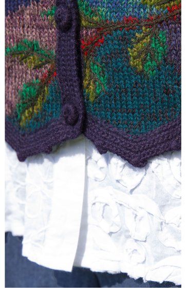 Strickweste in dunkellila mit farbigen Mustern in grün, rot und blau, Knöpfe in derselben Farbe, sechseckiger Saum, darüber weiße Bluse mit Rüschen und Volants