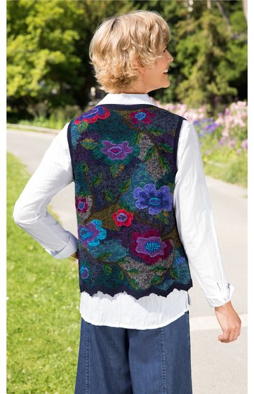Ärmellose Weste mit buntem Blumenmuster in Blau, Rot, Grün auf dunklem Grund, gerader Schnitt, an einer stehenden Person von hinten, weiße Bluse mit Kragen darunter, weit geschnittene blaue Hose
