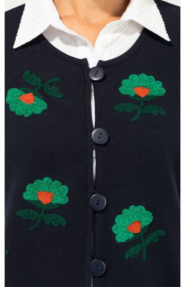 Dunkelblaue Strickjacke mit grünen gestickten Blumen mit orangenem Zentrum, Rundhalsausschnitt, vier schwarze Knöpfe, weißes Hemd mit spitzem Kragen darunter, Vorderseite sichtbar