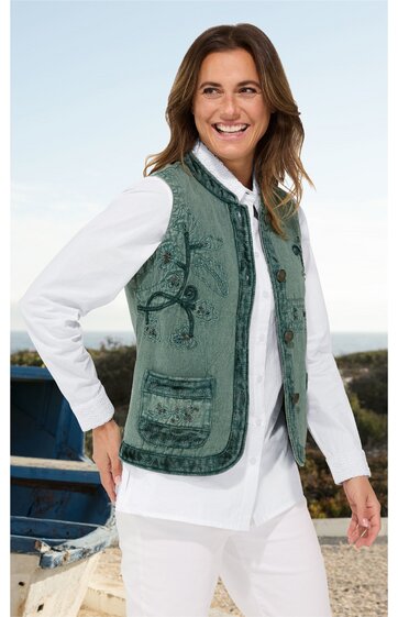 Damenweste in mittlerem Olivgrün mit floralem Muster, Stehkragen, durchgehender Reißverschluss, zwei aufgesetzte Taschen, locker sitzend, getragen von Frau in weißer Bluse und weißer Hose, stehend und lächelnd