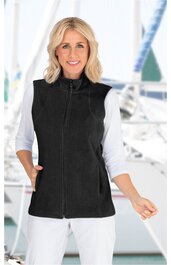 Schwarze ärmellose Weste mit hohem Stehkragen, durchgehendem Reißverschluss, zwei seitlichen Taschen, getragen von lächelnder stehender Frau in weißem Dreiviertelarmshirt vor maritimem Hintergrund