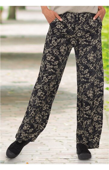 Schwarze Hose mit beigem floralen Muster, weite Beine, elastischer Bund, zwei Taschen, getragen von stehender Person mit Händen in den Taschen
