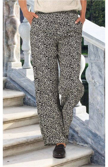 Locker sitzende schwarze Hose mit weißem abstraktem Linienmuster, gerade geschnitten, Seitentaschen, getragen von stehender Person mit Händen in den Taschen