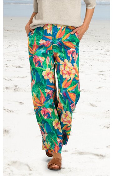 Helle Damenhose mit tropischem Blumenmuster in Grün, Orange, Gelb und Rosa, lockere Passform, elastischer Bund, lange Beine, getragen von stehender Person am Strand