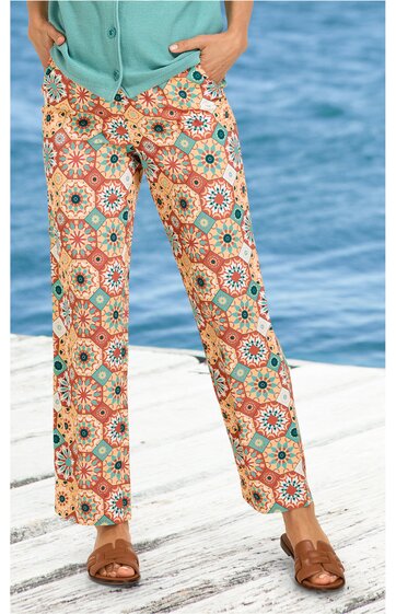 Hose mit weitem Bein, hellbeige Grundfarbe, buntes geometrisches Muster mit orange, blau und grün, gerade geschnitten, getragen von stehender Person, kombiniert mit türkisfarbenem Oberteil und braunen Sandalen
