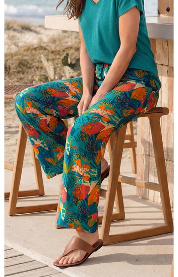 Weite, locker sitzende Hose mit farbigem Blumenmuster in Orange und Blau auf grünem Grund, getragen von sitzender Person, Kombiniert mit türkisem Kurzarmshirt und braunen Zehentrennern