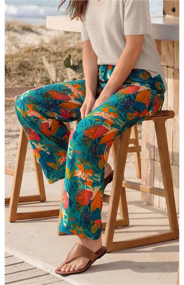 Hose in Türkis mit buntem Blumenmuster, locker geschnitten, lang, getragen von sitzender Person, kombiniert mit hellgrauem Kurzarmshirt und braunen Zehensandalen