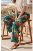 Hose in Türkis mit buntem Blumenmuster, locker geschnitten, lang, getragen von sitzender Person, kombiniert mit hellgrauem Kurzarmshirt und braunen Zehensandalen