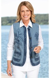 Ärmellose Weste in blau mit floralen Stickereien, Knopfleiste vorne, zwei aufgesetzte Taschen, über weißem Langarmhemd getragen, vorne offene Körperhaltung, lächelnde stehende Frau vor Gewässerkulisse